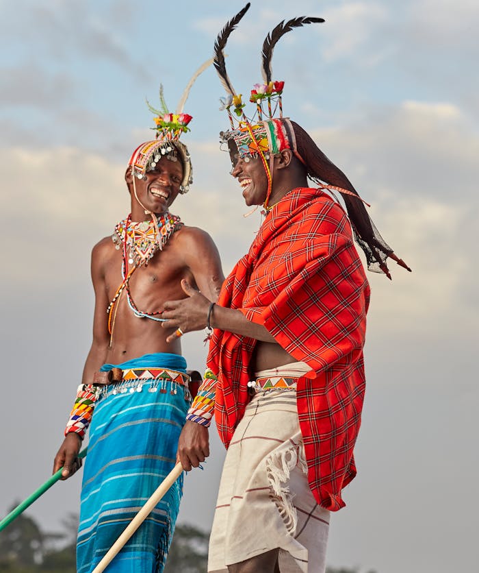 Two Maasai men in traditional attire smiling outdoors under a cloudy sky, embodying cultural heritage.